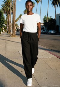 Frau geht auf einem sonnigen Bürgersteig entlang, der mit Palmen gesäumt ist, trägt ein weißes T-Shirt, schwarze Hose und weiße Turnschuhe, die Hände in den Taschen.