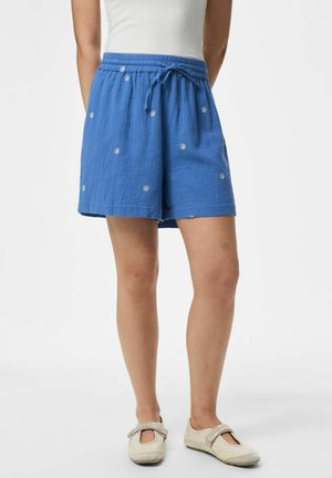 Personne portant un short bleu à cordon orné de petits motifs de fleurs blanches, un haut blanc et des chaussures Mary Jane beiges, debout devant un fond uni.
