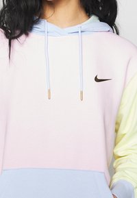 Mikina s kapucí v pastelově růžové, modré a žluté barvě. Obsahuje malé černé logo Nike a na koncích zlaté šňůrky. Jemný design s blokováním barev.