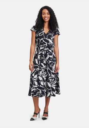 Schwarz-weißer Midi-Kleid mit floralem Muster, kurzen Ärmeln und einem Bindegürtel. Modell trägt schwarz-weiße High-Heels.
