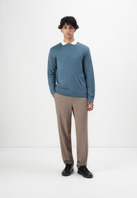 Jonge man die staat in een blauwe gebreide trui over een witte overhemd, bruine broek en zwarte sneakers, tegen een eenvoudige witte achtergrond.