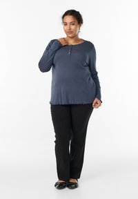 Marineblaues Langarmoberteil mit Knopfleiste, fransigem Saum, kombiniert mit schwarzen, geraden Hosen und schwarzen flachen Schuhen mit Verzierungen.