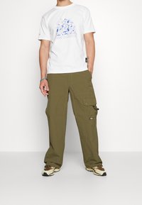 Vit t-shirt med blå grafisk design i kombination med olivgröna cargobyxor med fickor och en logotypapplikation. Bruna och beige sneakers.