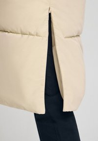 Beżowy pikowany płaszcz z bocznym suwakiem częściowo rozpiętym, ukazującym czarne spodnie pod spodem.