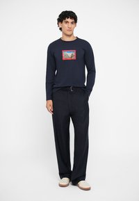 Navy long-sleeve T-shirt with a rectangular mountain graphic, paired with navy pinstriped trousers and beige sneakers.