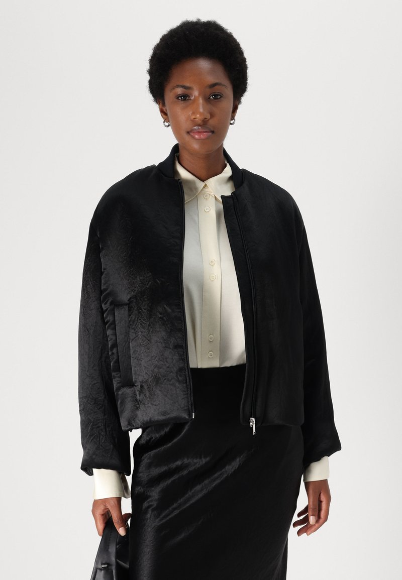 Veste bomber noire avec une finition texturée, fermeture éclair et col côtelé, associée à une chemise à boutons de couleur claire et une jupe noire élégante.