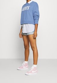 Sudadera azul recortada con el logo blanco de Levi's, superpuesta sobre una camiseta de rayas blanca y azul, combinada con pantalones cortos de mezclilla y zapatillas altas rosas de Nike.