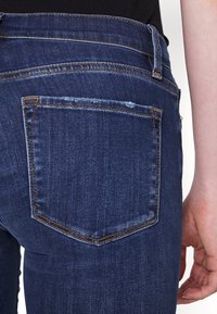 Jeans de denim azul oscuro con un bolsillo trasero, con costuras en contraste amarillas y un ligero borde desgastado en el bolsillo.