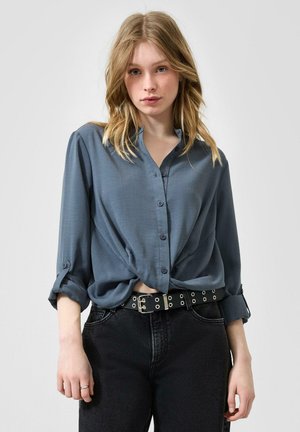 Jeune femme aux cheveux blonds portant une chemise bleue ample boutonnée avec les manches retroussées et un pantalon noir avec une ceinture noire avec des œillets argentés.