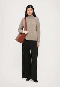 Frau in einem beigen, kabelgestrickten Rollkragenpullover, schwarzen weit geschnittenen Hosen, schwarzen Schuhen und mit einer großen braunen Schultertasche.
