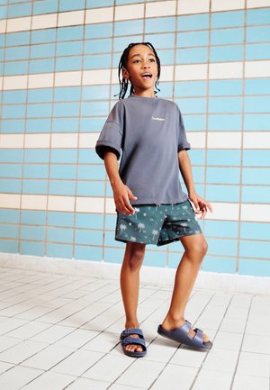 Otrok s pletenicami, ki nosi sivo preveliko majico, zelene kratke hlače z vzorcem palme in mornarsko modre sandale, stoji ob modrem opečnem zidu.