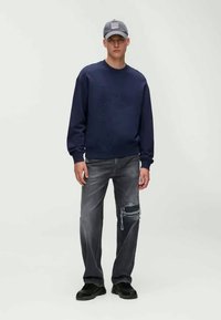 Navy sweatshirt with textured front design, paired with faded grey distressed jeans. The model wears a grey cap and black shoes.