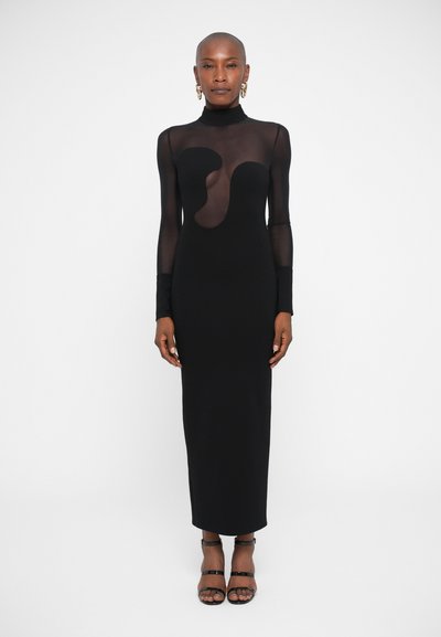 Robe noire à manches longues avec une silhouette ajustée, présentant une section en maille transparente et un design découpé unique. Des escarpins noirs simples complètent le look.