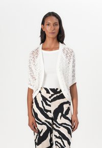 Cardigan en tricot ouvert blanc avec un motif en zigzag, porté par-dessus un top blanc. Associé à un pantalon large à rayures zèbres beige et noir.