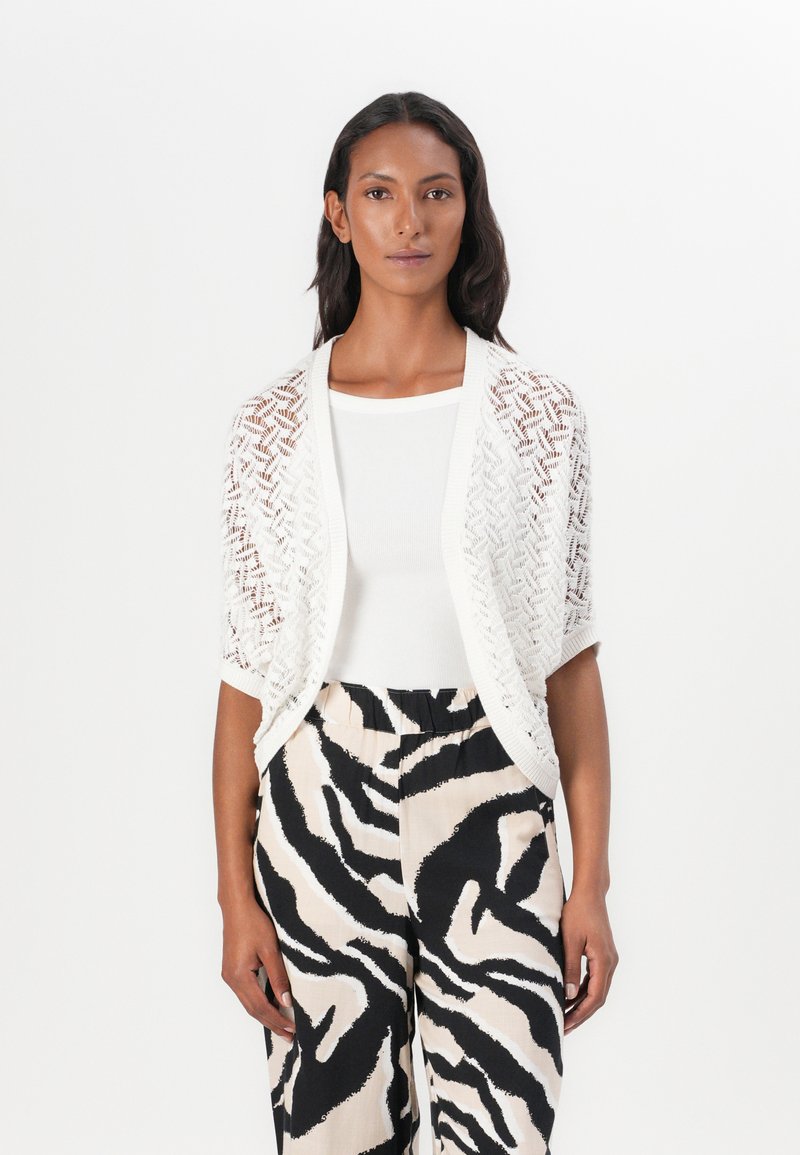 Cardigan en tricot ouvert blanc avec un motif en zigzag, porté par-dessus un top blanc. Associé à un pantalon large à rayures zèbres beige et noir.