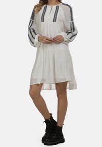 Biała sukienka z długimi rękawami, plisowanym dołem i niebieskimi wyszywanymi akcentami. Posiada guziki i wiązanie pod szyją. Czarne buty na sznurowadła.