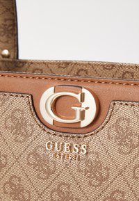 Borsa a tracolla marrone con texture, dotata di accessori in oro, motivo goffrato e chiusura a logo circolare. "GUESS EST 1981" in lettere dorate sul fronte.