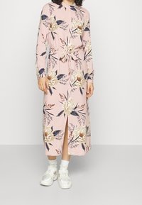 Lungo abito rosa con stampa floreale, maniche lunghe, chiusura a bottoni, vita cintrata, spacco frontale e tessuto comodo. Indossato con sneaker bianche.
