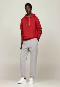 Ung man som står iförd en röd hoodie, ljusgrå sweatpants och vita sneakers mot en enfärgad bakgrund.