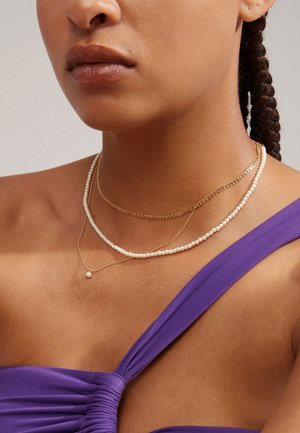 Gold and pearl necklaces layered over a purple top; features a delicate chain, multiple strands, and a simple pearl pendant.