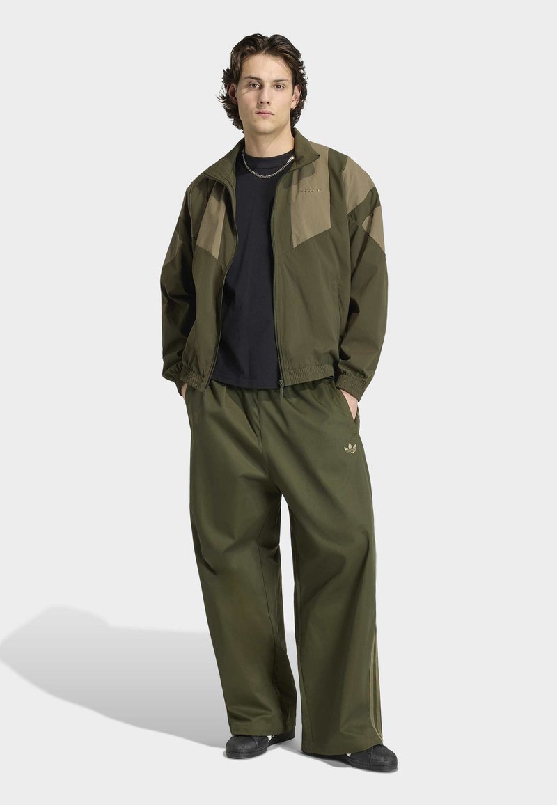 Jeune homme debout les mains dans les poches, portant une veste et un pantalon de sport vert olive, un t-shirt noir et des chaussures noires sur fond clair.