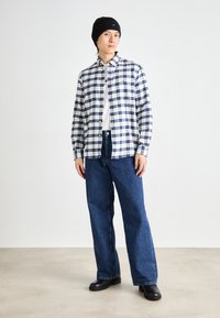 Karierter Hemd aus Baumwolle in Weiß und Marineblau; darunter ein weißes T-Shirt. Weite blaue Jeans und eine schwarze Mütze. Schwarze Stiefel vervollständigen das Outfit.