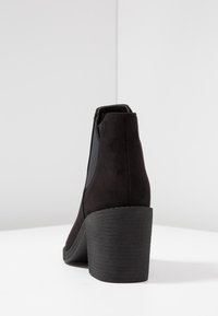 Bottine noire en suede doux avec un bout arrondi et un talon empilé. Dispose de panneaux latéraux en élasthanne pour un enfilage facile.