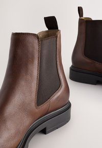 Hnedé kožené členkové topánky s elastickými bočnými panelmi, textúrovaným povrchom a čiernou gumovou podrážkou. Majú jazýček na ľahké obúvanie.