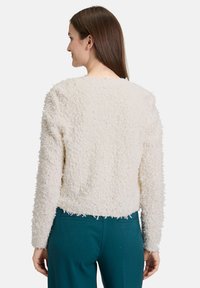 Femme aux cheveux bruns lisses portant un pull en crème texturé à manches longues et un pantalon teal, faisant face à un fond blanc.