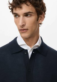Granatowy sweter z dzianiny z kołnierzykiem polo, zapinany na guziki, o lekkiej teksturze, noszony na białej koszuli z kołnierzykiem.