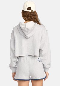 Vrouw gezien van achteren, gekleed in een lichtgrijze cropped hoodie, bijpassende shorts met marineblauwe afwerking en een witte honkbalpet.