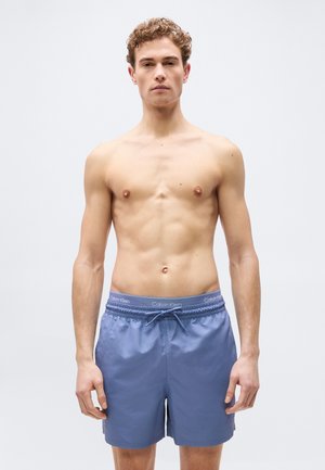 Muž bez košile s kudrnatými vlasy, oblečený v světle modrých plavkách Calvin Klein, stojící před jednobarevným světle šedým pozadím.