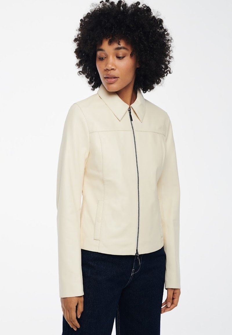 Vrouw met krullend haar in een crèmekleurige jas met een rits aan de voorkant en een kraag, gecombineerd met donkerblauwe broek, staande tegen een witte achtergrond.