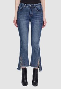 Jeans in denim blu con vita aderente, fondo svasato e dettagli scalfiti alle caviglie, caratterizzati da accenti colorati e finitura con bordo grezzo.