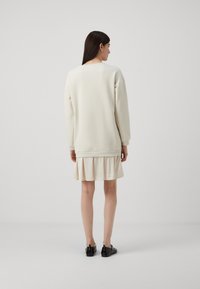 Sweatshirt crema con polsini a coste, indossato sopra un vestito ruffled di un crema chiaro; abbinato a scarpe nere. Design minimalista con texture liscia.