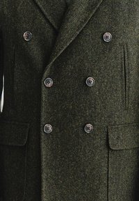 Dwurzędowy płaszcz wełniany w ciemnozielony wzór jodełki. Posiada sześć guzików, dwie przednie kieszenie oraz wyłogi z wcięciami.