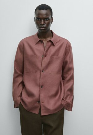 Camicia bordeaux a maniche lunghe con bottoni, dotata di due tasche frontali e una texture morbida, abbinata a pantaloni marrone scuro. Design minimalista e sfondo neutro.