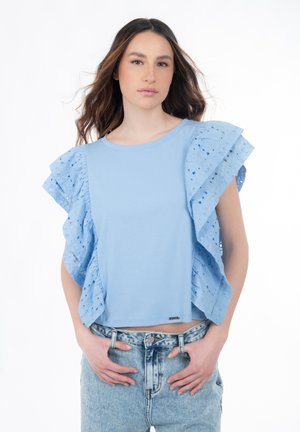 Jonge vrouw met golvend bruin haar, gekleed in een lichtblauw topje met geruite oogjesmouwen en licht gespoelde hooggetailleerde spijkerbroek, met de handen in de zakken.