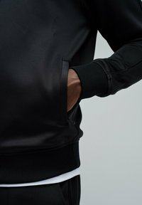 Chaqueta atlética negra con una textura suave, puños acanalados y un bolsillo lateral. Superpuesta sobre una camiseta blanca en el dobladillo.