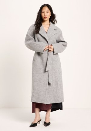 Woman wearing a long light gray belted coat over a burgundy dress and black pointed heeled shoes, standing on a plain light background.