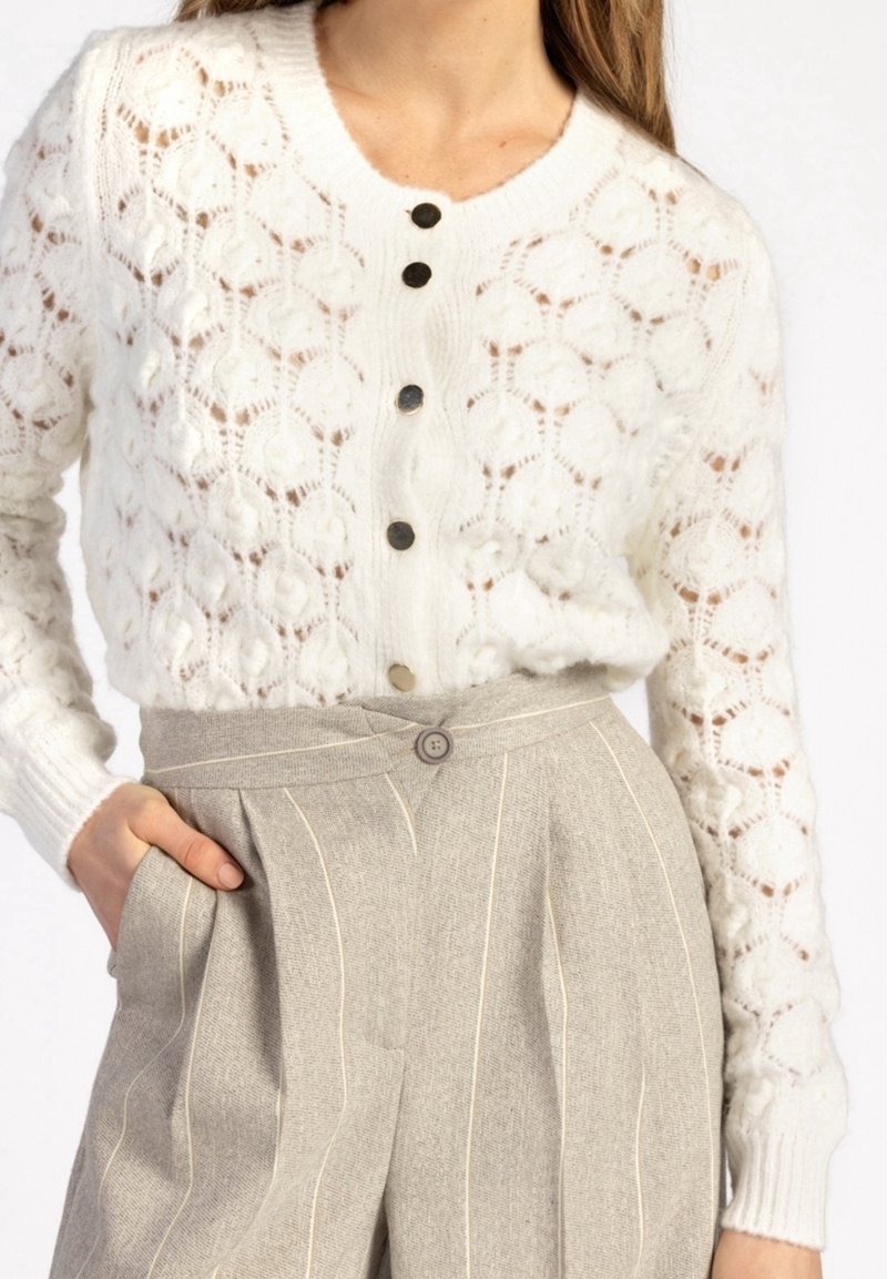 Cardigan en maille blanc à manches longues avec motif texturé et boutons noirs, associé à un pantalon beige à taille haute à fines rayures.