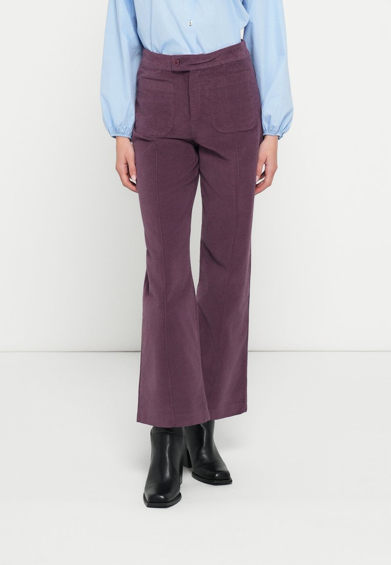 Lila Schlaghosen mit strukturierter Oberfläche, ausgestattet mit Fronttaschen und einem Knopfverschluss, kombiniert mit einem langärmligen hellblauen Hemd.