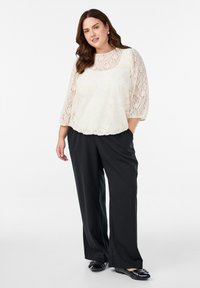 Sieviete ar caurspīdīgu krēma krāsas mežģīņu blūzi pār atbilstošu topu, melnām platām biksēm un melnām dzīvokļu kurpēm ar sudraba studiem, stāv ar vienu roku kabatā.