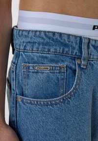 A világoskék farmernadrág hátulján egy ezüst logóval ellátott zseb és narancssárga cérna díszíti. Fent egy fehér derékpánt látható, szürke csíkokkal.