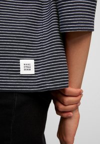 Person, der ein navyblaues und weiß gestreiftes Hemd mit einem "Marc O'Polo Denim"-Label trägt und sein anderes Handgelenk hinter dem Rücken hält.