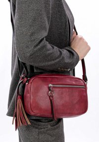 Borsa a tracolla rossa in pelle con una texture liscia, dotata di una tasca frontale con zip, di una tracolla nera e di dettagli decorativi in frange.