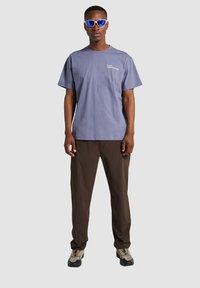 Fioletowa bawełniana koszulka z małym białym logo, w połączeniu z brązowymi spodniami typu cargo i szarymi butami do wędrówki z czarnymi podeszwami i sznurowadłami.