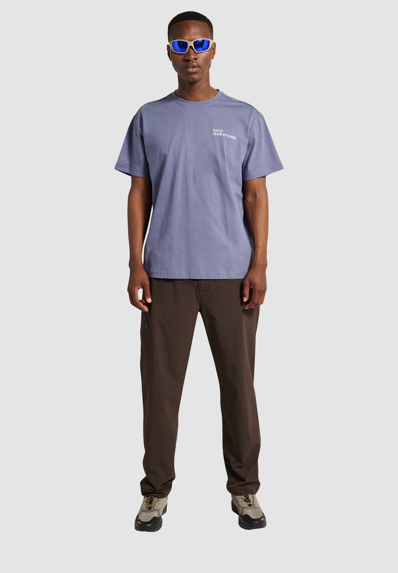 Mężczyzna w niebieskich okularach przeciwsłonecznych, szarej koszulce z nadrukiem, ciemnobrązowych spodniach i beżowych butach turystycznych stoi na jednolitym jasnym tle.