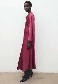 Robe longue et fluide de couleur bordeaux avec un décolleté drapé et de larges manches en forme de cloche. Associée à des bottines noires à la cheville.