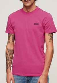 Maglietta di cotone fucsia con maniche corte, scollo rotondo e piccolo logo navy. Presenta tatuaggi visibili sulle braccia e una vestibilità comoda.